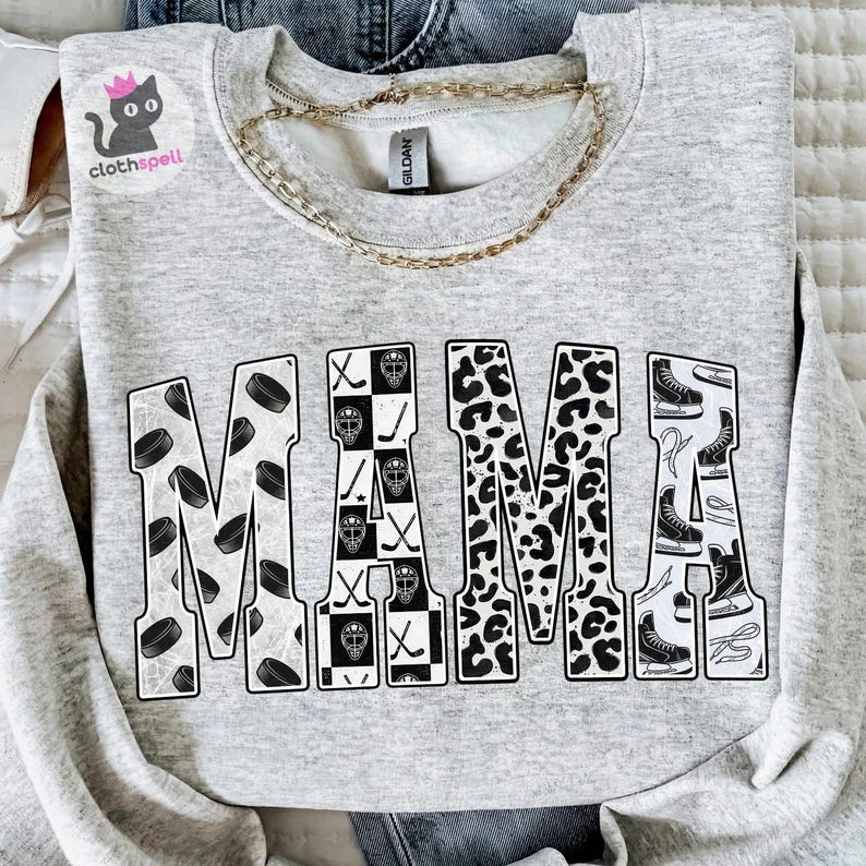 May include: A light grey crewneck sweatshirt featuring the word MAMA in large block letters. The letters are filled with black and white patterns including hockey pucks, a checkered hockey mask design, leopard print, and ice skates.