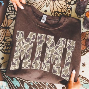 May include: A brown sweatshirt with the name "MIMI" in large, block letters. The letters are filled with a camouflage pattern in shades of brown, tan, and gray. The sweatshirt is being held up by a person.