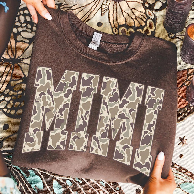May include: A brown sweatshirt with the name "MIMI" in large, block letters. The letters are filled with a camouflage pattern in shades of brown, tan, and gray. The sweatshirt is being held up by a person.