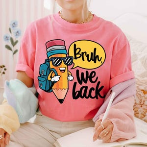 May include: Pink t-shirt featuring a cartoon pencil wearing sunglasses and a backpack. The pencil is in front of a yellow speech bubble with the words "Bruh we back" in black. The shirt is a casual style.