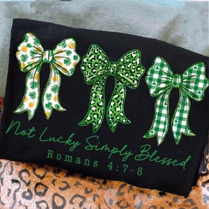 May include: Black t-shirt featuring three green bows with different patterns: shamrocks and gold coins, leopard print, and green gingham. The text "Not Lucky Simply Blessed" and "Romans 4:7-8" are printed in green.