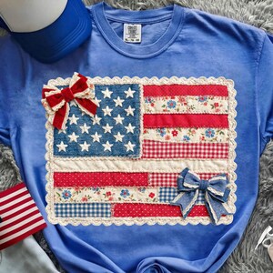 Puede incluir: Una camiseta azul claro con un dise&ntilde;o de bandera americana de patchwork. La bandera incluye estrellas azules vaqueras, rayas rojas y blancas y patrones florales y de cuadros. Dos lazos decorativos est&aacute;n unidos a la bandera.
