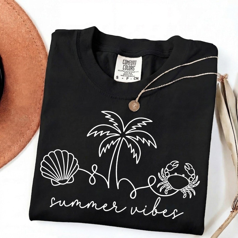May include: Black t-shirt with a white graphic of a seashell, palm tree, and crab, with the words "summer vibes" in a script font. A gold necklace with a pendant is on the shirt. The shirt has a Comfort Colors tag.