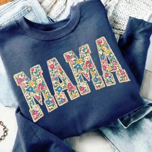 K&ouml;nnte beinhalten: Marineblaues Sweatshirt mit dem Wort "MAMA" in gro&szlig;en Buchstaben mit Blumenmuster. Die Buchstaben sind goldfarben umrandet. Das Sweatshirt liegt gefaltet auf einer Jeans.