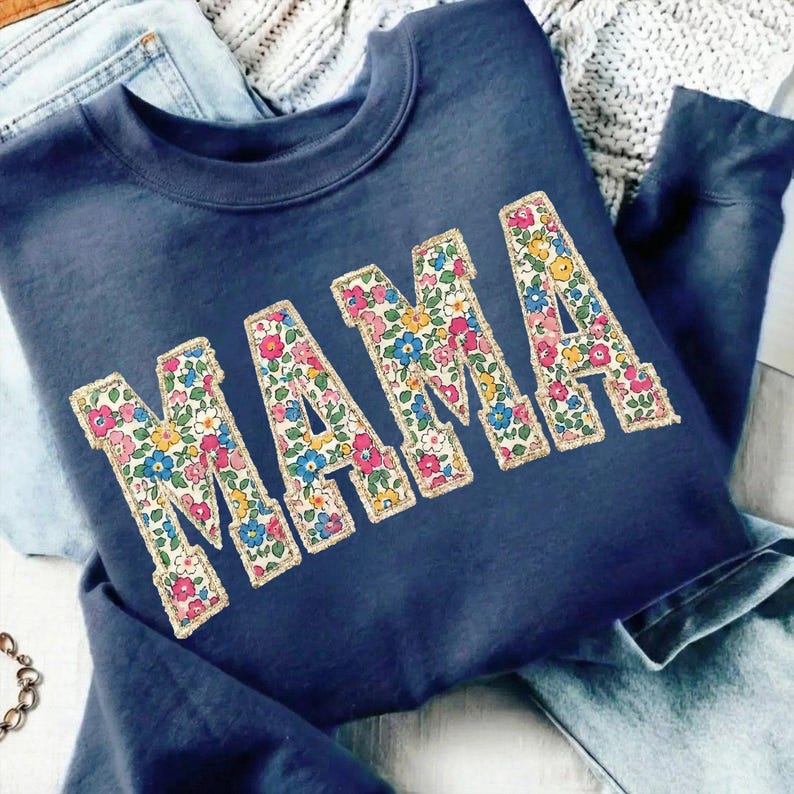 K&ouml;nnte beinhalten: Marineblaues Sweatshirt mit dem Wort "MAMA" in gro&szlig;en Buchstaben mit Blumenmuster. Die Buchstaben sind goldfarben umrandet. Das Sweatshirt liegt gefaltet auf einer Jeans.
