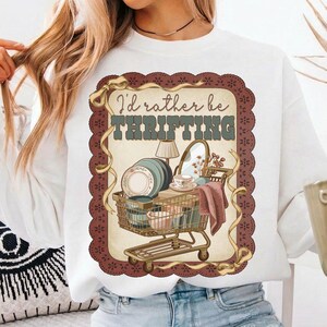 May include: White sweatshirt with a graphic featuring a shopping cart filled with vintage items like plates, a lamp, and a mirror. The text "I'd rather be THRIFTING" is displayed above the cart, framed by a decorative border.