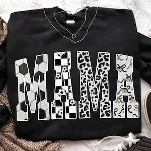 May include: Black sweatshirt with the word "MAMA" in large, white letters. The letters are decorated with soccer ball patterns, checkered patterns, leopard print, and bows. A gold necklace is draped across the top.