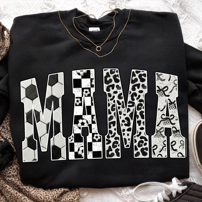 May include: Black sweatshirt with the word "MAMA" in large, white letters. The letters are decorated with soccer ball patterns, checkered patterns, leopard print, and bows. A gold necklace is draped across the top.