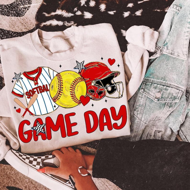 May include: A cream-coloured sweatshirt featuring a softball design. The graphic includes a softball jersey, a yellow softball, a red helmet, and the words "GAME DAY" in red. The design is accented with hearts and stars.