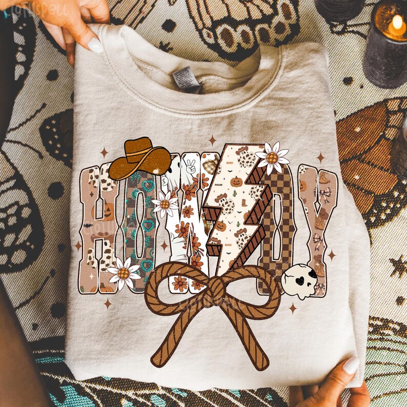 May include: Beige sweatshirt with the word "HALLOWAY" in a western-themed design. The design includes a cowboy hat, flowers, a lightning bolt, and a rope bow. The sweatshirt is folded.