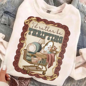 May include: White sweatshirt with a graphic featuring a shopping cart filled with vintage items like plates, a mirror, and a lamp. The text "I'd rather be thrifting" is displayed above the cart in a vintage style.