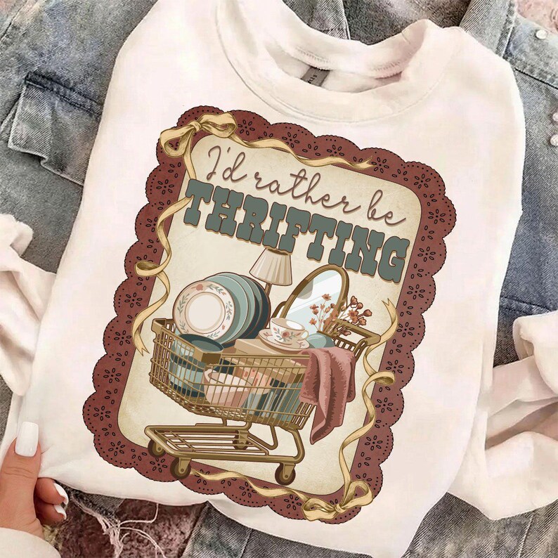 May include: White sweatshirt with a graphic featuring a shopping cart filled with vintage items like plates, a mirror, and a lamp. The text "I'd rather be thrifting" is displayed above the cart in a vintage style.