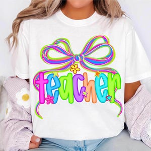 May include: White t-shirt with a colorful bow and the word "teacher" in vibrant, multi-colored bubble letters. The design includes small flowers and stars, creating a cheerful and playful aesthetic. A perfect gift for teachers.