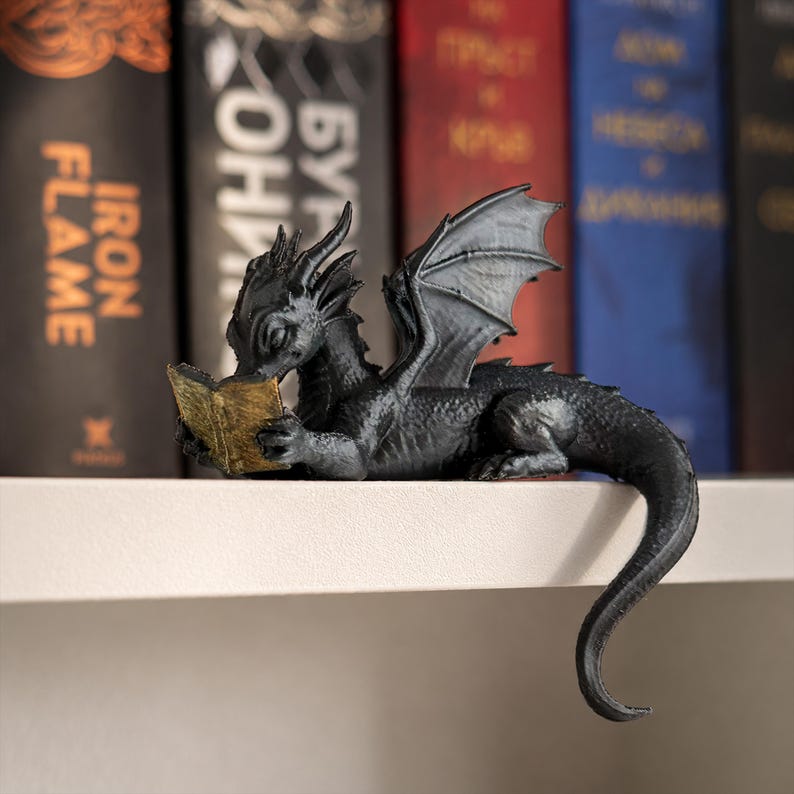 Detailed black dragon figurine reading a miniature gold book on a white bookshelf with softly blurred red, blue and black fantasy novels behind it.