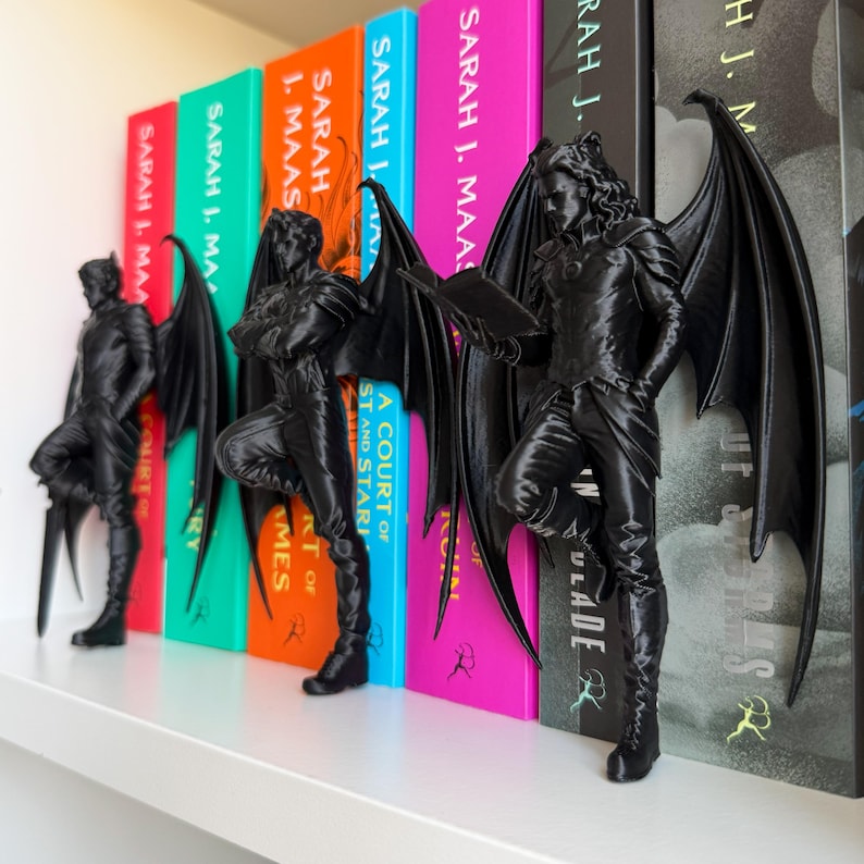 Pu&ograve; includere: Segnalibri neri a forma di figure alate su una mensola bianca accanto a libri colorati. I fermalibri sono ricchi di dettagli. I libri hanno titoli di Sarah J. Maas.