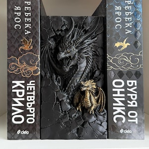 Könnte beinhalten: Eine schwarze Buchstütze mit einer großen, detaillierten Drachenskulptur und einer kleineren goldenen Drachenfigur davor, platziert zwischen zwei schwarzen Büchern mit kyrillischem Text, goldenen Drachensymbolen und Wolkenmustern auf den Buchrücken.