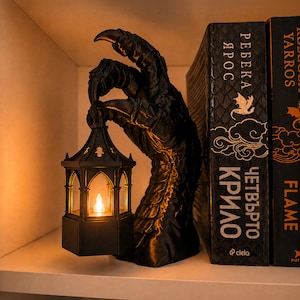 May include: A black, textured dragon claw sculpture holding a small, hexagonal lantern with a glowing, warm-toned light inside. The sculpture is positioned next to two books with black covers and gold lettering, including the text "ЧЕТВЪРТО КРИЛО".