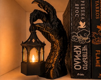 May include: A black, textured dragon claw sculpture holding a small, hexagonal lantern with a glowing, warm-toned light inside. The sculpture is positioned next to two books with black covers and gold lettering, including the text "ЧЕТВЪРТО КРИЛО".
