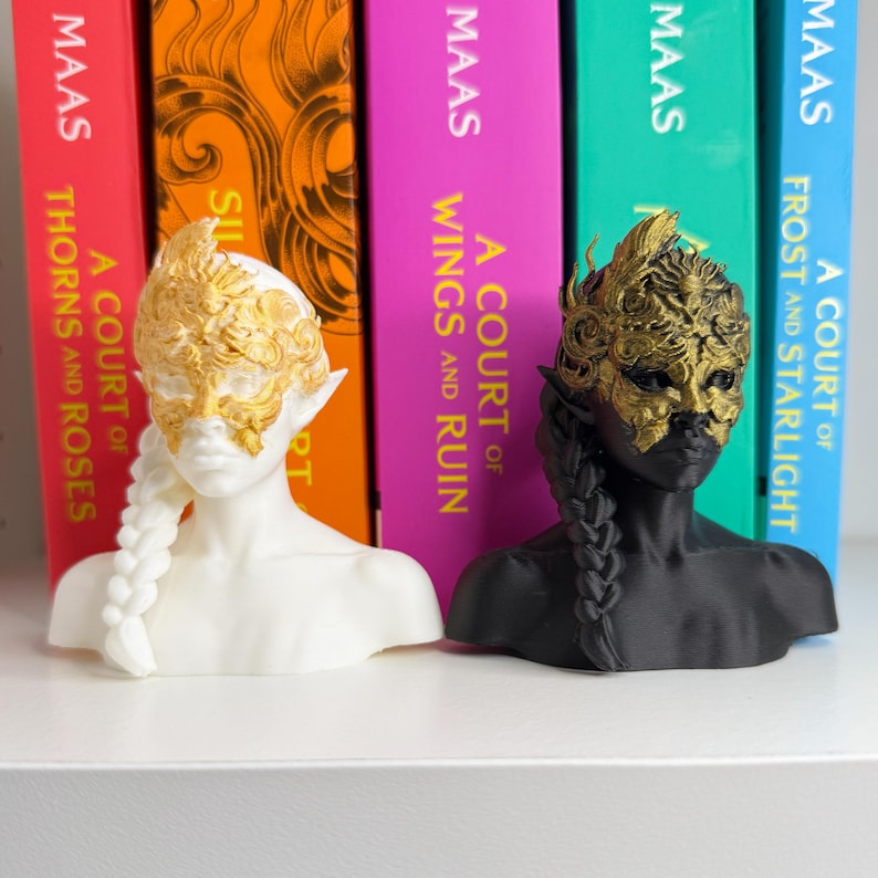May include: Two busts of a female figure with pointed ears and a braided hairstyle. One is white with a gold mask and headpiece, the other is black with a gold mask and headpiece. Books with colorful spines are in the background.
