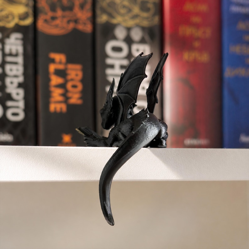 Rear view of a black reading dragon bookshelf figurine showing its raised wings and long curved tail hanging over the white shelf edge.