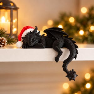 Op de afbeelding: Een zwart drakenbeeldje met gestructureerde schubben en vleugels, met een kleine rood-witte kerstmuts op. De draak is afgebeeld in een slapende houding, met het lichaam rustend op een plat oppervlak en de lange, stekelige staart over de rand.