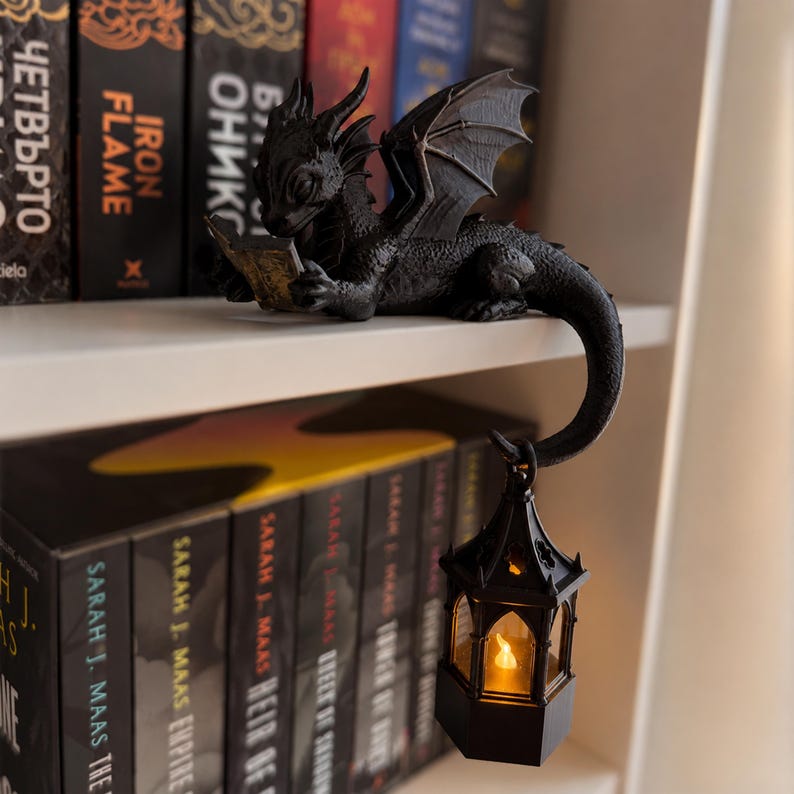 3D printed dragon reading a book on the edge of a shelf with a warm glowing LED lantern below.