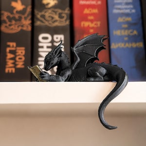 Close side view of a black 3D printed reading dragon with raised wings, gold book and long tail hanging over the edge of a bookshelf.