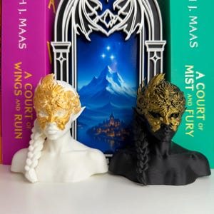May include: Two busts of figures with pointed ears and braided hair, one white and one black. Both wear ornate gold masks. Behind them are books with the titles "A Court of Wings and Ruin" and "A Court of Mist and Fury".