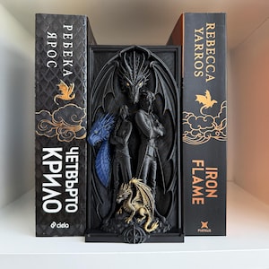 Pode incluir: Um suporte para livros preto com um relevo esculpido de duas figuras diante de um grande dragão, com um dragão dourado menor na base e uma cabeça de dragão azul à esquerda. O suporte está posicionado entre dois livros intitulados Fourth Wing e Iron Flame.