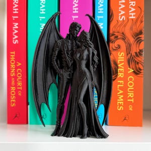 Peut inclure: Une figurine noire imprimée en 3D représentant deux personnages ailés debout ensemble. Le personnage masculin a de grandes ailes de chauve-souris et porte une veste à col, tandis que le personnage féminin a de longs cheveux fluides et une robe longue.