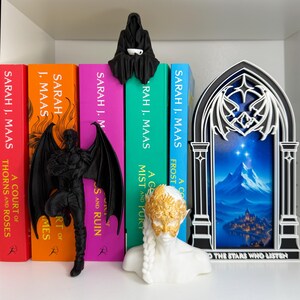 May include: A shelf display featuring fantasy-themed bookends and figurines. Includes a black winged figure, a white bust with a gold mask, and a black cloaked figure holding a cup. Books with colorful spines are visible, with titles by Sarah J. Maas.