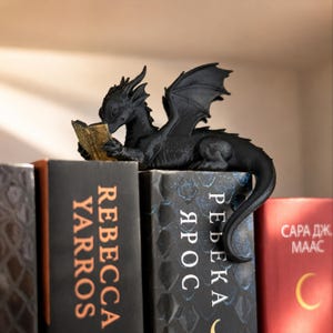 Könnte beinhalten: Eine schwarze Drachenfigur mit texturierten Schuppen und Flügeln, die auf einem Buchrücken sitzt. Der Drache hält ein kleines, offenes Buch mit goldfarbenen Seiten. Die Figur steht zwischen Büchern mit Text, darunter Rebecca Yarros.