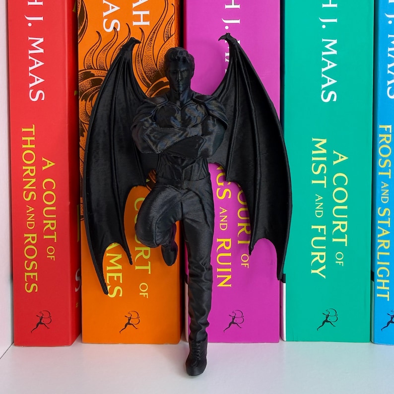 May include: A black figurine of a winged humanoid figure, leaning against a row of colorful books. The books have titles like "A Court of Thorns and Roses" and "A Court of Mist and Fury."
