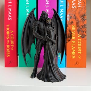 May include: A black figurine of two winged figures standing together, with one figure wearing a gold-colored mask. The figures are positioned in front of four colorful book spines, including titles by Sarah J. Maas such as A Court of Thorns and Roses.