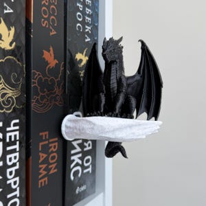 May include: A black dragon figurine perched on a white, textured, hand-shaped bookend. The dragon has detailed scales and large, spread wings. The bookend is positioned against a row of books, including one with the text Iron Flame visible.