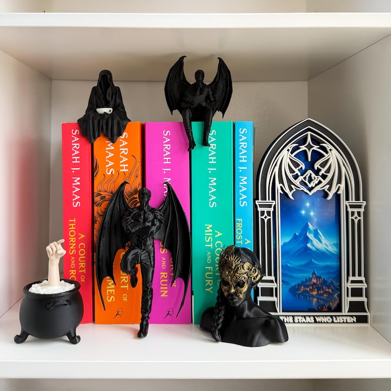 May include: A shelf displays fantasy-themed decor and books. Black figurines of winged creatures and a cauldron with a hand are arranged with colorful book spines. The books are by Sarah J. Maas.