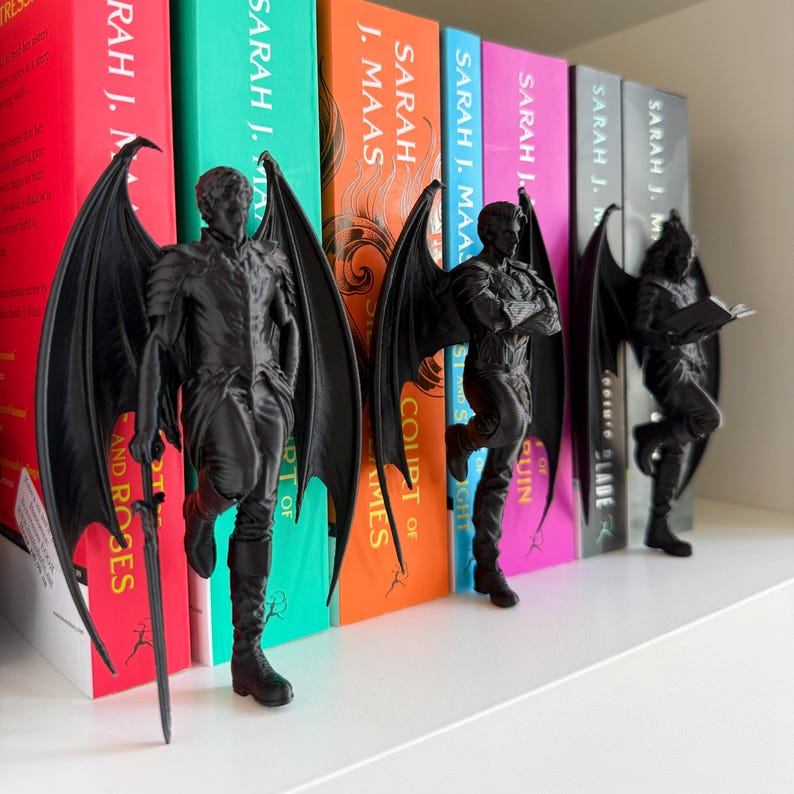 Pu&ograve; includere: Statuette nere di figure alate, ognuna in posa diversa, appoggiate a una fila di libri colorati. Una figura tiene una spada, un'altra ha le braccia conserte e l'ultima legge un libro. I libri sono di Sarah J. Maas.