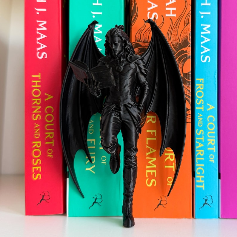 Pu&ograve; includere: Una statuetta nera di una figura alata, che legge un libro, si trova di fronte a una fila di libri colorati. I libri hanno titoli come "A Court of Thorns and Roses" e "A Court of Frost and Starlight" di H.J. Maas.