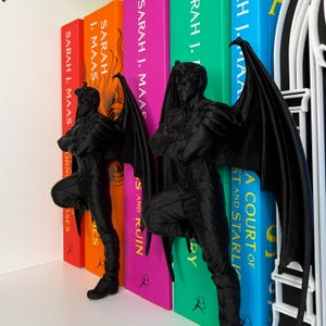 May include: Black bookends shaped like a winged humanoid figure, leaning against colorful book spines. The bookends are positioned between the books, adding a decorative and functional element to a bookshelf. The books have titles by Sarah J. Maas.
