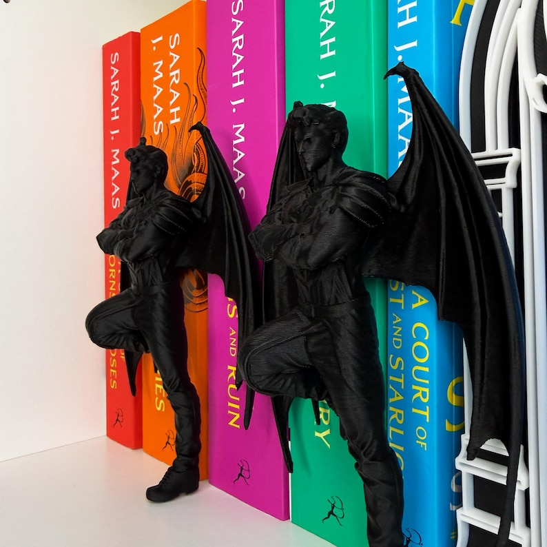 May include: Black bookends shaped like a winged humanoid figure, leaning against colorful book spines. The bookends are positioned between the books, adding a decorative and functional element to a bookshelf. The books have titles by Sarah J. Maas.