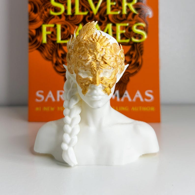 May include: A white bust of a figure with a gold mask and headpiece, and a long, white braid. The bust is in front of a book with the title "Silver Flames" on the cover.