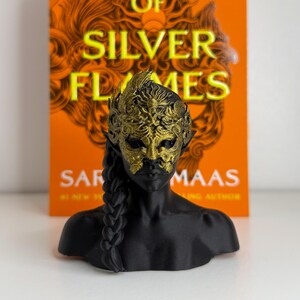 May include: A black bust sculpture of a person wearing a gold mask with intricate details. The sculpture has a braided hairstyle. Behind the bust is a book with the title "OF SILVER FLAMES" in large yellow letters on an orange background.