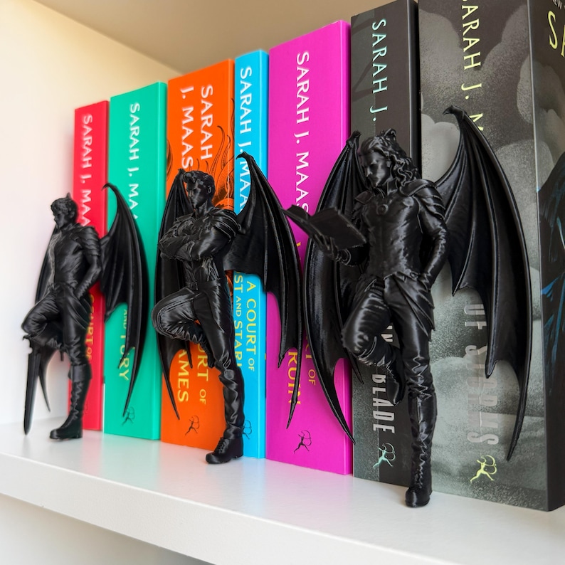 Pu&ograve; includere: Figure nere alate fungono da fermalibri su una mensola bianca, affiancando dorsi di libri colorati. Le figure sono dettagliate, una che tiene una spada e un'altra un libro. I libri sono di Sarah J. Maas.