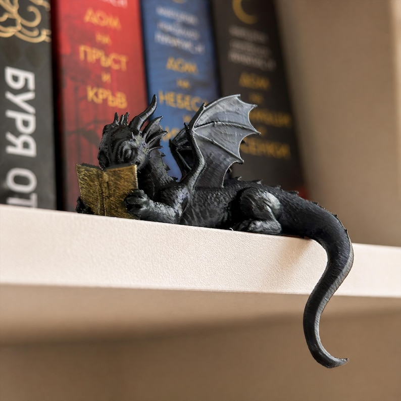 Black reading dragon bookshelf figurine holding a small gold book on a white shelf with softly blurred fantasy books in the background.