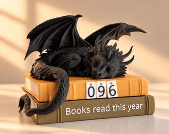 May include: A black dragon figurine with textured scales and spread wings resting on two stacked books. The top book is yellow with the number 096 displayed, and the bottom book is olive green with the text Books read this year.