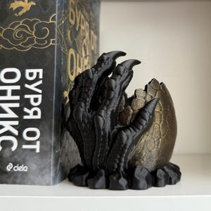 May include: A black 3D-printed dragon claw sculpture clutching a textured, metallic bronze-colored egg. The claw features detailed scales and sharp talons, resting on a rocky base. The object is positioned next to a book with Cyrillic text.