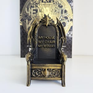 以下が含まれることがあります： 背もたれに「MY HOUSE MY CHAIR MY WOMAN」の文字が入った、黒と金のミニチュアの玉座。玉座には、ドラゴンの翼のようなアクセントや装飾的な彫刻など、装飾的なディテールが施されています。背景には金色の文字が入った本があります。