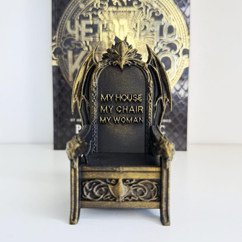 以下が含まれることがあります： 背もたれに「MY HOUSE MY CHAIR MY WOMAN」の文字が入った、黒と金のミニチュアの玉座。玉座には、ドラゴンの翼のようなアクセントや装飾的な彫刻など、装飾的なディテールが施されています。背景には金色の文字が入った本があります。