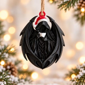 Könnte beinhalten: Ein schwarzer Hängeschmuck, der eine Kapuzenfigur mit großen fledermausartigen Flügeln zeigt, die eine rot-weiße Weihnachtsmütze trägt und eine kleine weiße Tasse hält. Der Schmuck ist an einer beigen Schnur befestigt und zeigt detaillierte Strukturen.