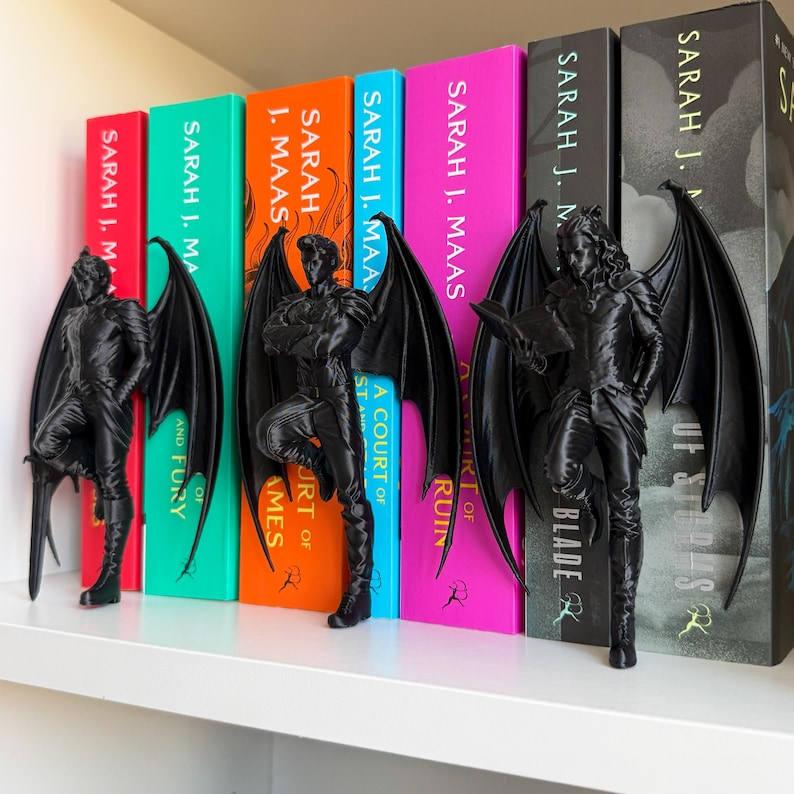 May include: Black, winged figure bookends stand beside colorful books with titles by Sarah J. Maas. The bookends are detailed sculptures of a character with large wings. The books have titles like "A Court of Thorns and Roses" and "A Court of Mist and Fury."